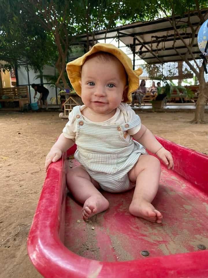 Adorable baby sitting on red outdoor play equipment in natural setting.