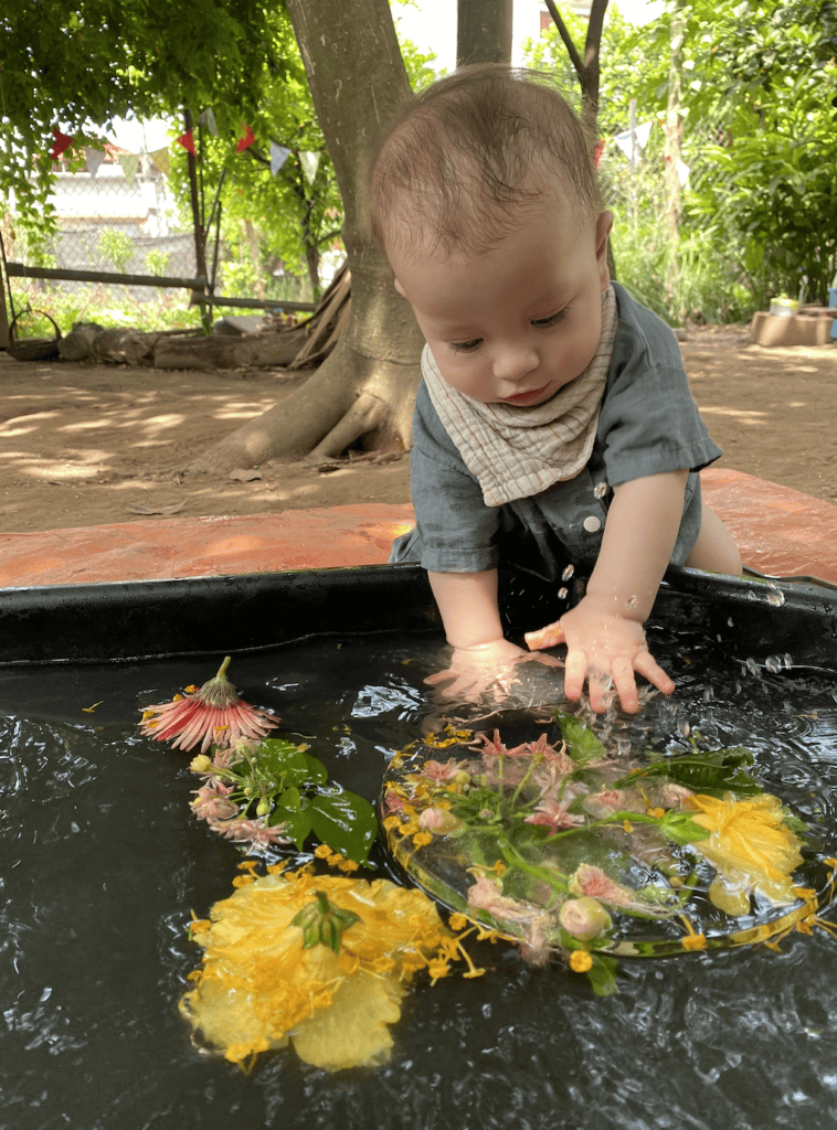 Baby playing with floating flowers in water at outdoor play area.